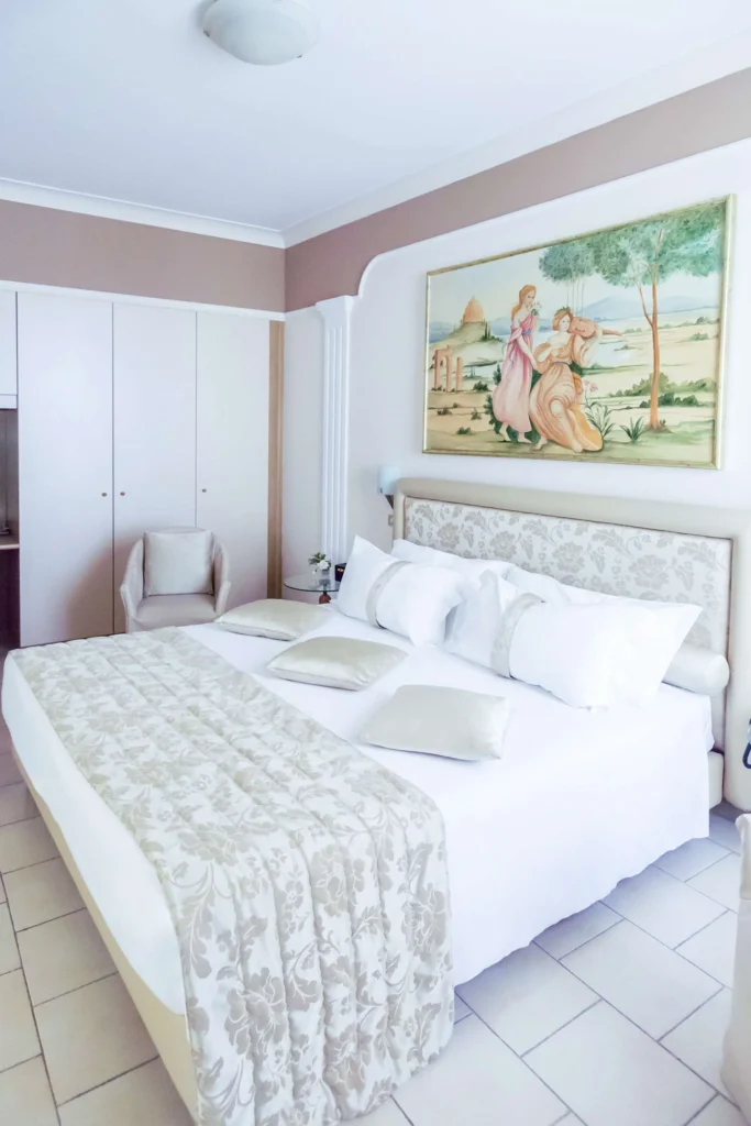 Nell'immagine la camera family dell'hotel. La stanza è occupata da un grande letto matrimoniale con runner beige e, accanto, una poltroncina beige dietro alla quale si vede un armadio a muro. Sopra il letto è appeso un quadro colorato.