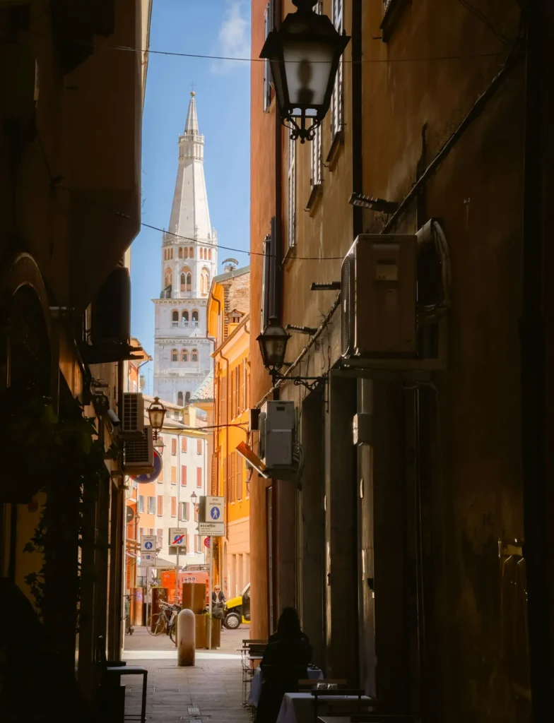 Nell'immagine si vede uno scorcio del centro storico di Modena con un vicoletto in ombra in primo piano e il campanile del Duomo illuminato sullo sfondo.